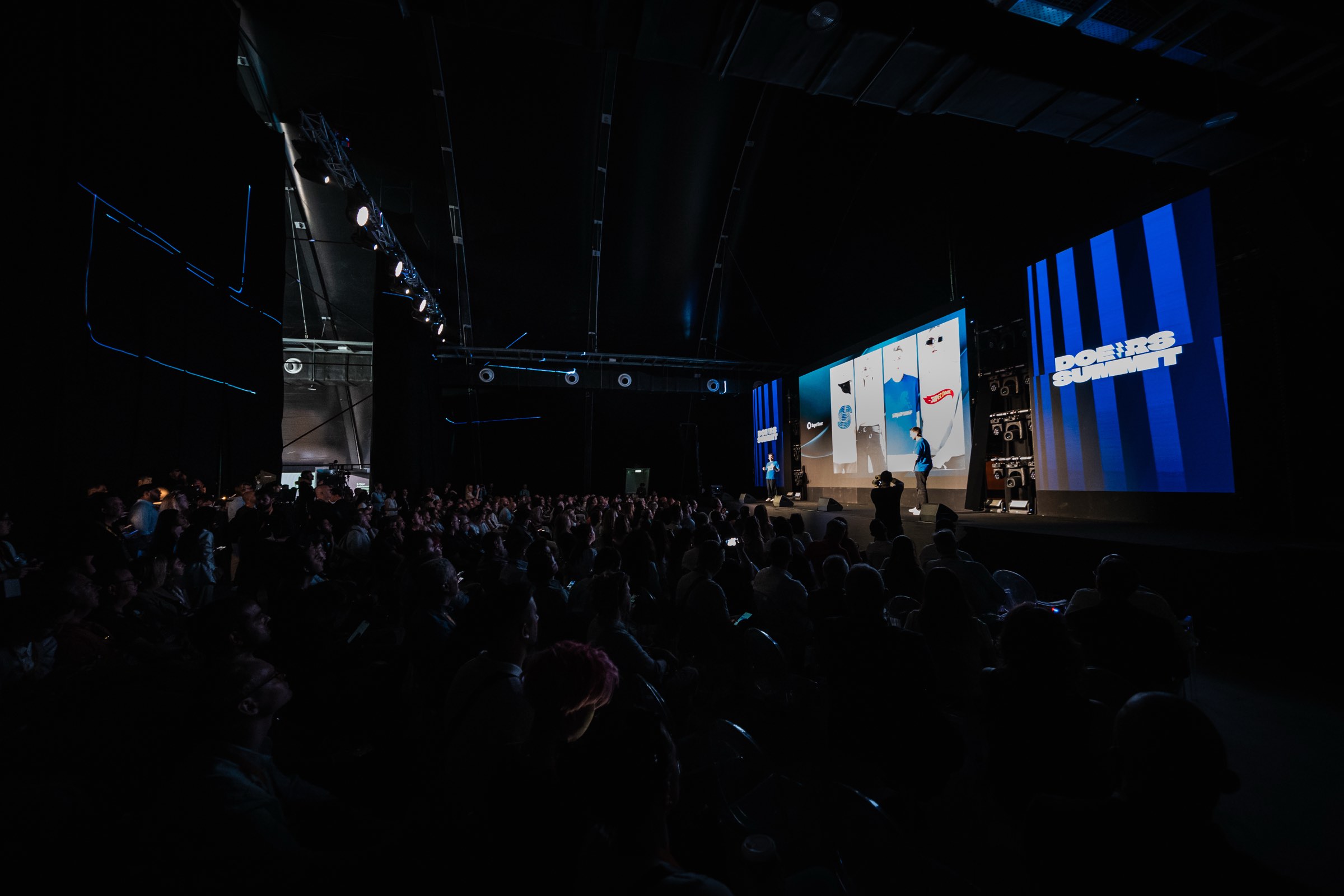 A full Doers Summit auditorium in the dark, facing a blue-lit stage and the Doers Summit screens