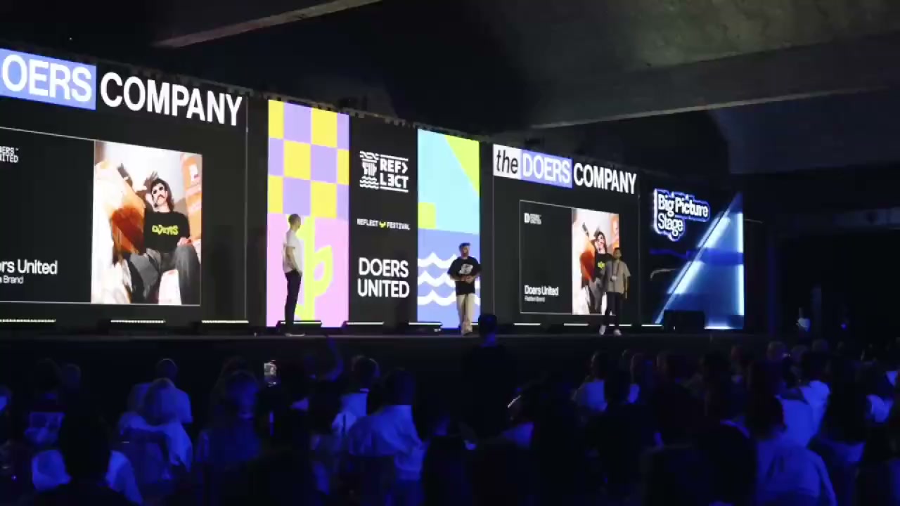 A blue-lit Doers Summit crowd facing a wall of Doers Company and Doers United screens