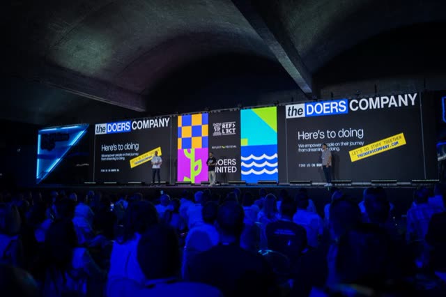 A packed hall facing a wide LED wall carrying The Doers Company branding