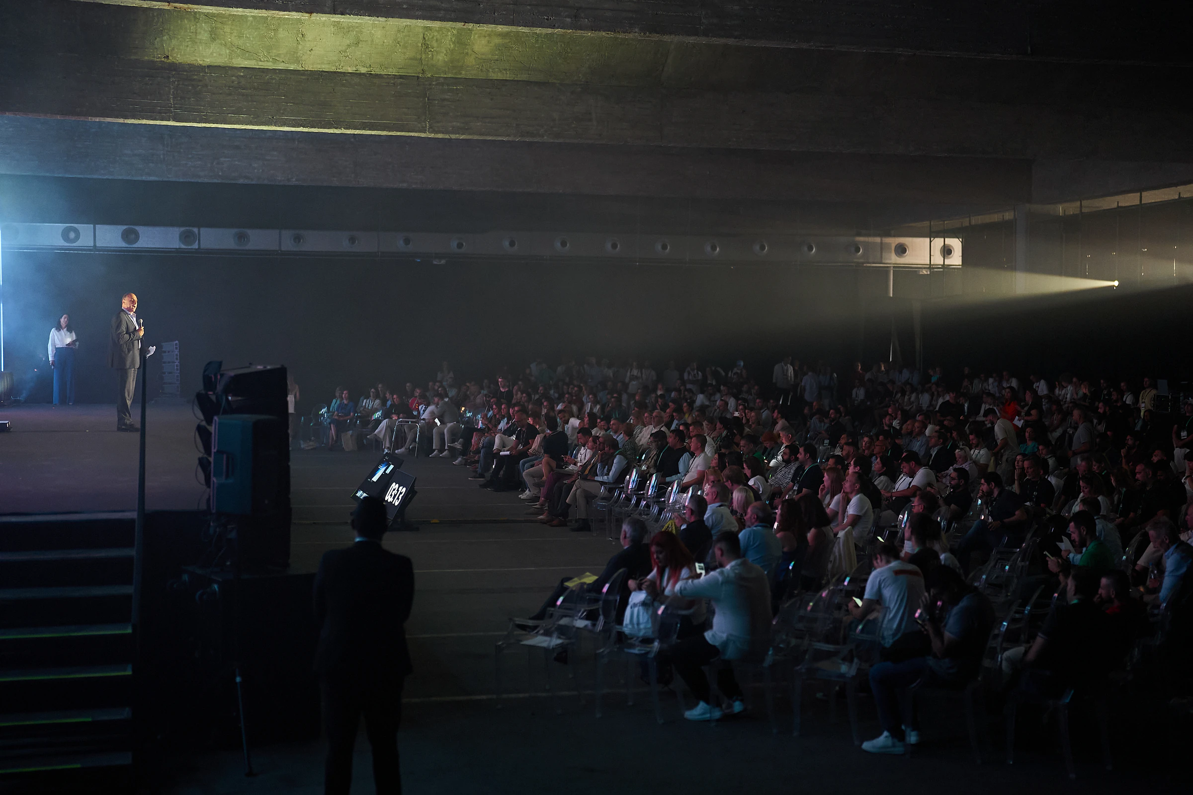 A speaker on a darkened stage addressing a full seated audience at Doers Summit